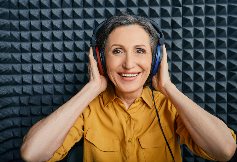 A smiling woman sits in a soundproof booth while wearing headphones and undergoing a series of hearing health tests.