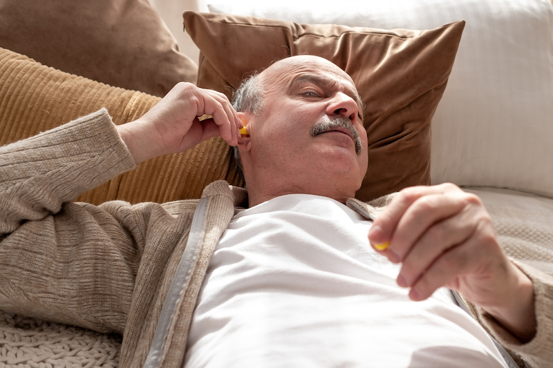 A man lies down on his bed, placing earplugs in his ears.