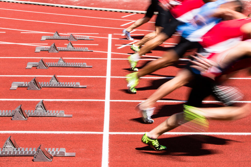 Blurred image of sprinters launching from the starting blocks on a red track during a race, capturing the intense motion and energy of the athletes.