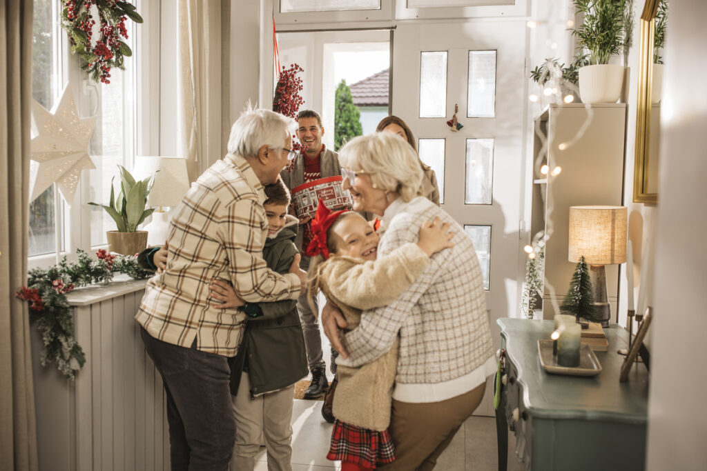 Grandparents warmly hugging their grandchildren during a festive holiday gathering with family entering through a decorated doorway.