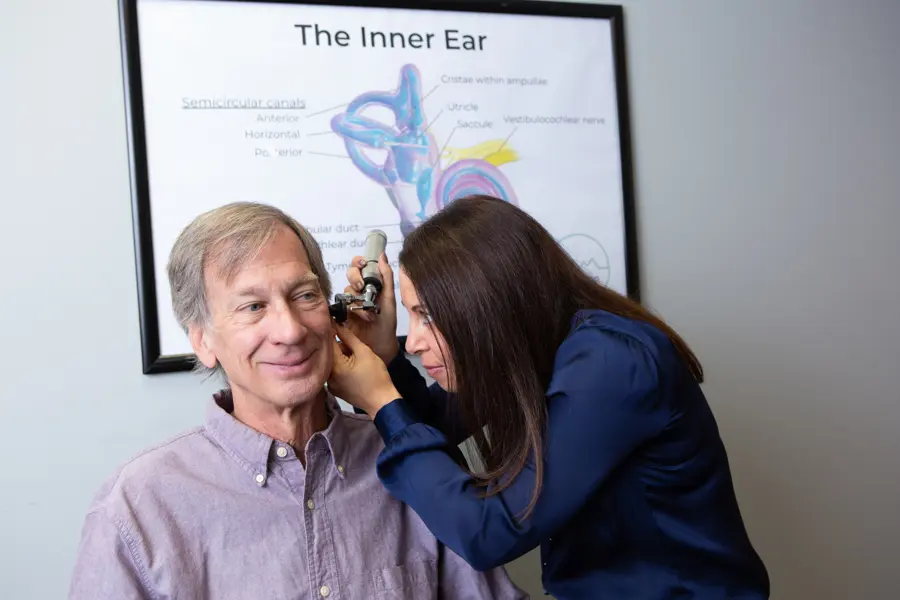 Dr. Meredith Wilken examines an older mans ear with an otoscope in a medical office. Behind them is a poster showing a diagram labeled The Inner Ear. Both appear calm and focused.