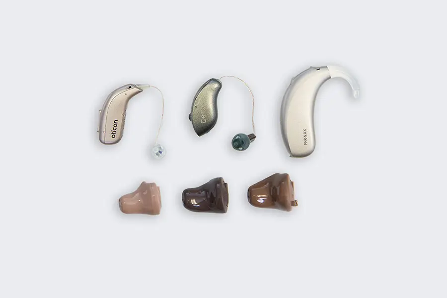 A collection of six hearing aids in various styles and colors, including behind-the-ear and in-the-ear models, arranged in two rows on a light gray background.