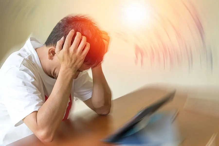 A person sitting at a table holds their head in their hands, appearing stressed or overwhelmed, with blurred, swirling light effects around their head suggesting confusion or anxiety. A tablet and papers are on the table.