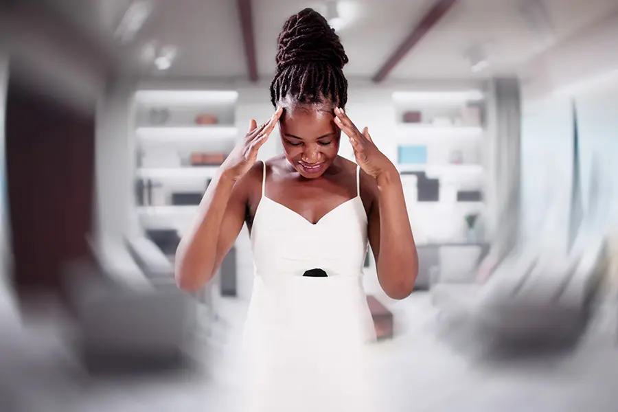 A woman in a white dress stands indoors, touching her temples and looking down, with a blurred background suggesting dizziness or disorientation.