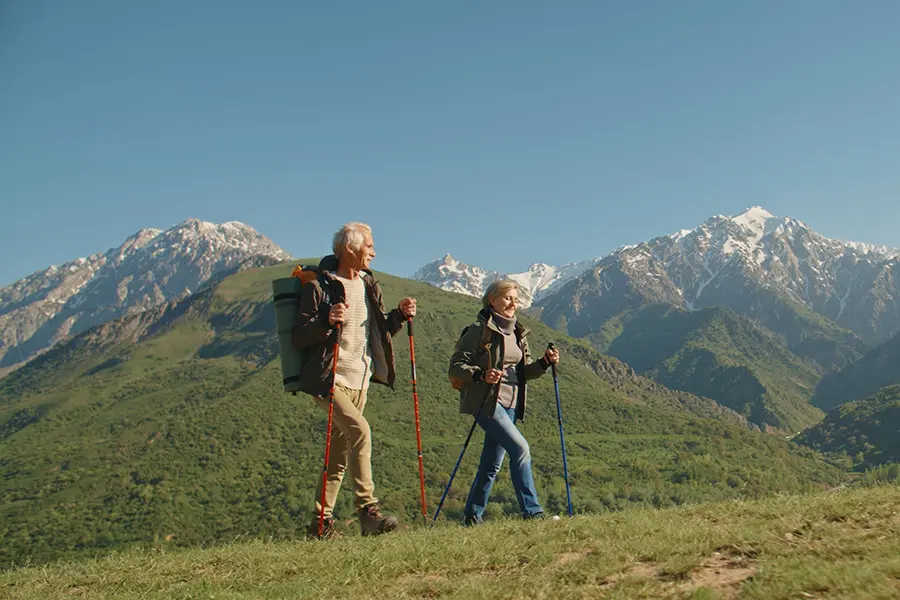 An older man and woman hike with trekking poles on a grassy hill, surrounded by green valleys and snow-capped mountains under a clear blue sky.