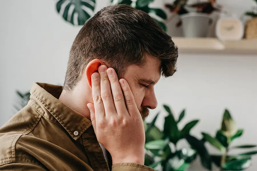 A man with short brown hair and a beard holds his hand to his ear, appearing to be in discomfort or pain. He is indoors with green plants and shelves in the background.