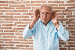Older man in glasses cupping his hands behind his ears to hear, as when hearing aids sound uneven, from Colorado Ear Care near Denver, CO